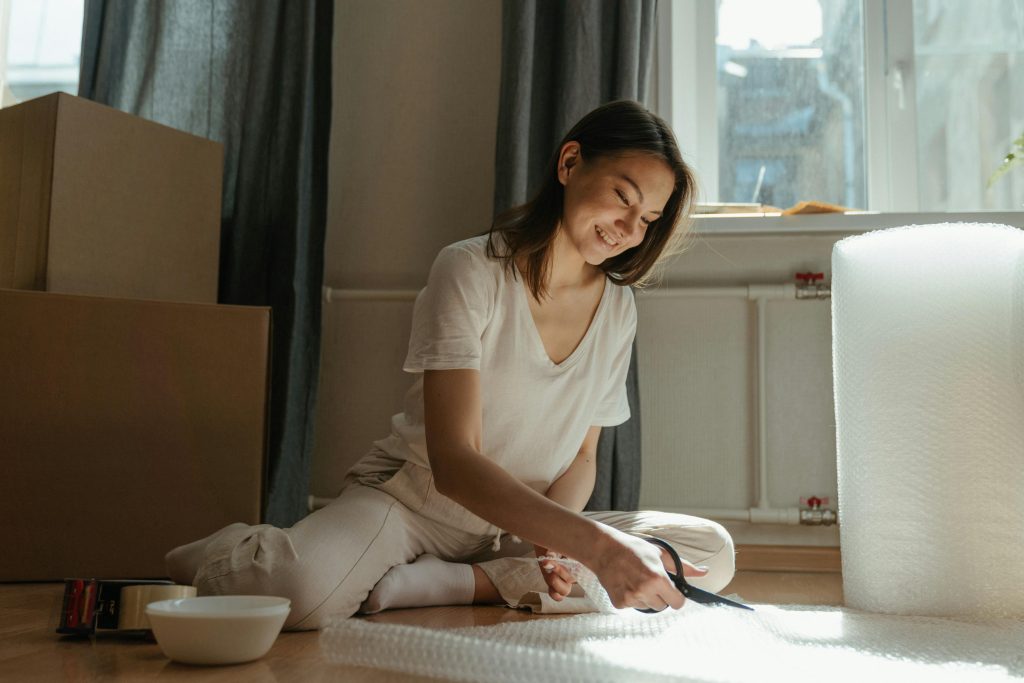 Young woman packing belongings with bubble wrap by a window, preparing for a move.